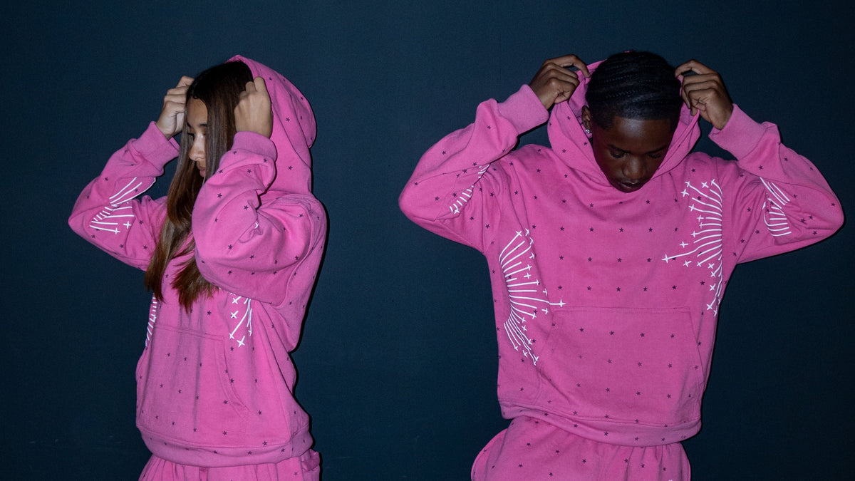 Two models wearing matching bright pink hooded sweatshirt and jogger sets with white starburst prints and small dark polka dots, posed against a dark navy background.